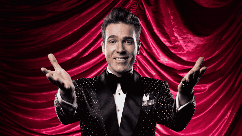 A smiling man in a sparkly black tuxedo with a bow tie stands in front of dramatic red curtains, extending both arms forward in a welcoming gesture, capturing the excitement of a Broadway performance.