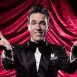 A smiling man in a sparkly black tuxedo with a bow tie stands in front of dramatic red curtains, extending both arms forward in a welcoming gesture, capturing the excitement of a Broadway performance.