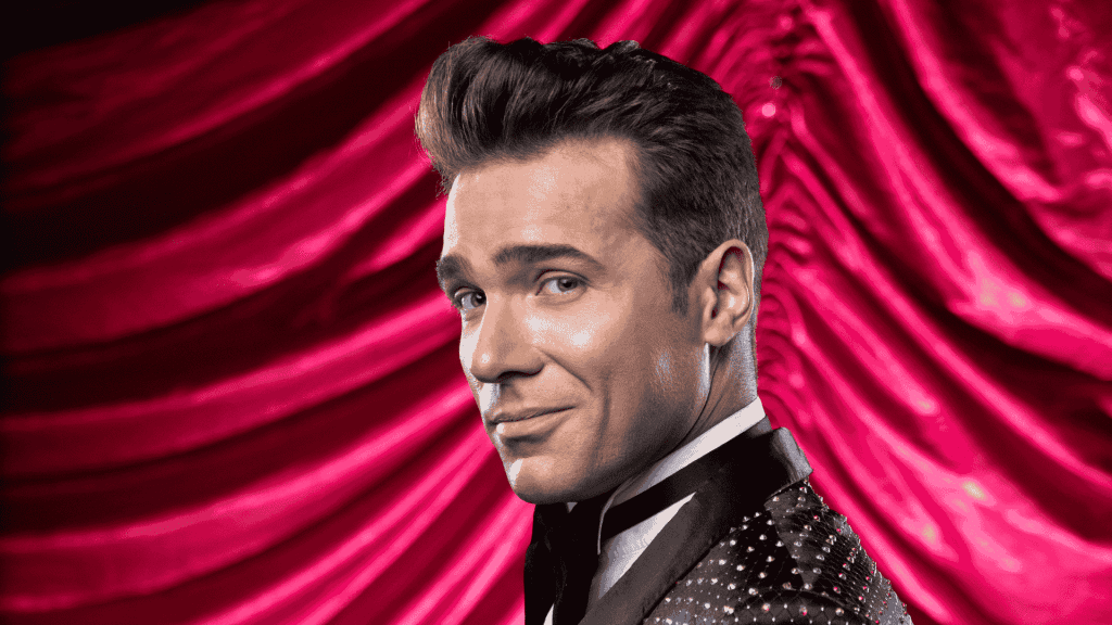 Jonathan Roxmouth, in a sparkling black tuxedo, smiles confidently over his shoulder against a dramatic, red velvet curtain backdrop.