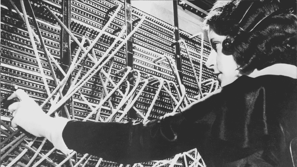 A woman operates a vintage telephone switchboard, connecting calls by inserting cables into various ports. Amid a tangle of wires, her focused demeanor recalls the playful coordination found in a bustling schoolyard.