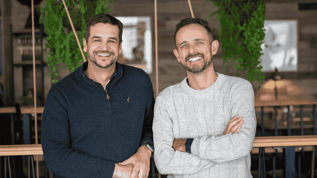 Two men smiling, standing side by side indoors with greenery and wooden decor in the background—perhaps sharing a memorable expat explore experience. One wears a dark sweater, the other a light sweater with arms crossed.