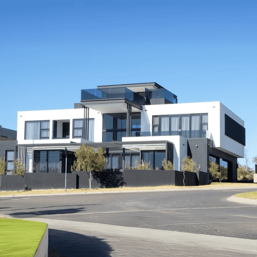 A modern, multi-story house with white and black exterior, large windows, and balconies, situated on a sunny day with clear blue sky and minimal landscaping in front.