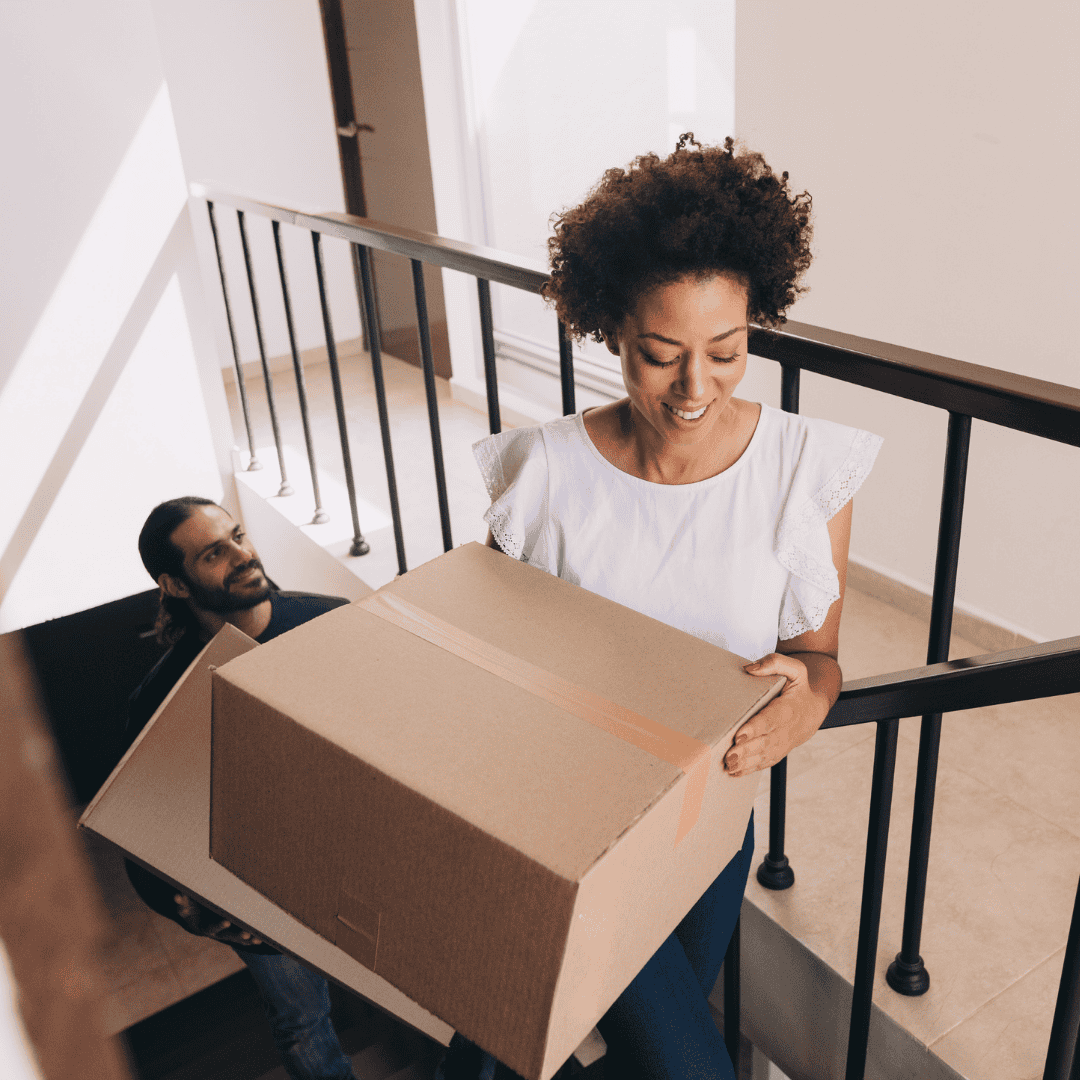 A woman smiles while carrying a large cardboard box up a staircase, with a man behind her also holding a box. The setting appears to be a residential building or apartment complex.