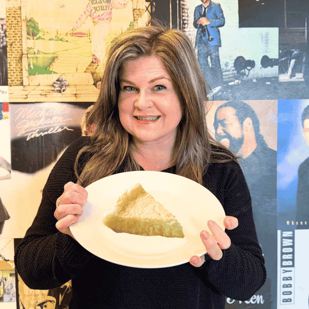 A smiling woman with long brown hair holds a white plate with a slice of pie in front of a colorful wall decorated with various album covers and music posters.