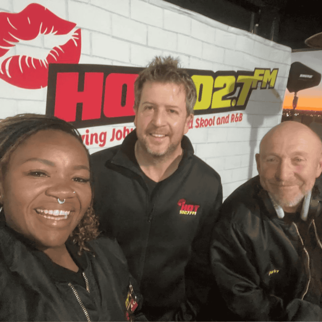 Three people smiling and posing together in front of a wall with a Hot 102 FM radio station logo and a red lips graphic. All are wearing black jackets, and headphones are around one person’s neck.