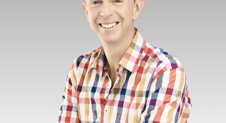 A smiling man with short hair wearing a colorful plaid shirt stands with his arms crossed against a light gray gradient background, exuding confidence and the energy of a hot business professional.