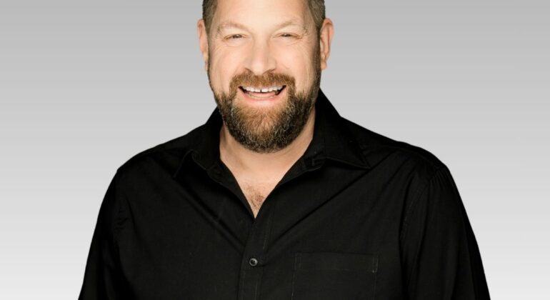 Patrick, a bearded man wearing a black button-up shirt, smiles at the camera in front of a light gray gradient background.