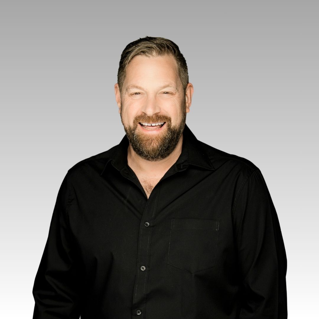 Patrick, a bearded man wearing a black button-up shirt, smiles at the camera in front of a light gray gradient background.