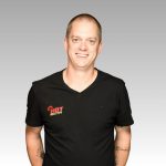 A smiling man with short hair wears a black HOT 102.7 FM t-shirt and stands against a plain, light gray background. His arms hang relaxed by his sides, and he has visible tattoos on both forearms.