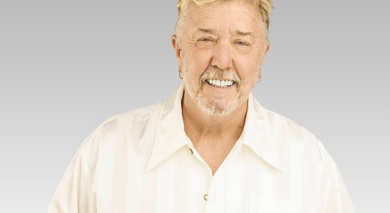 Smiling older man with short blond hair and a trimmed beard, wearing a white collared shirt, standing against a plain gray background.