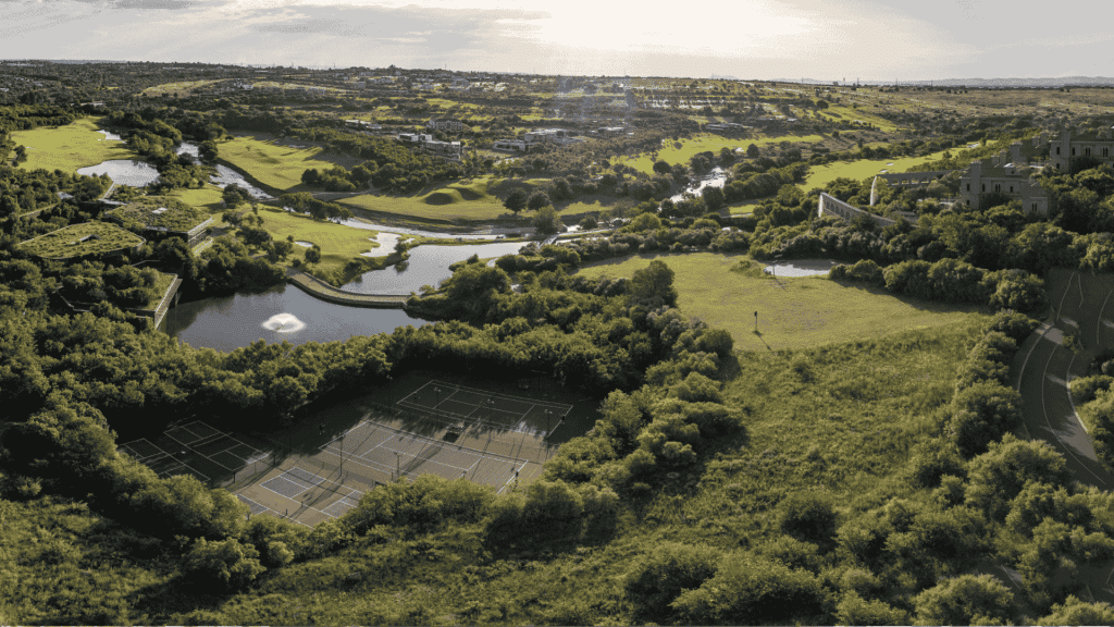Aerial view of a lush green landscape featuring tennis courts, winding rivers, ponds, open fields, and scattered trees under a bright sky.