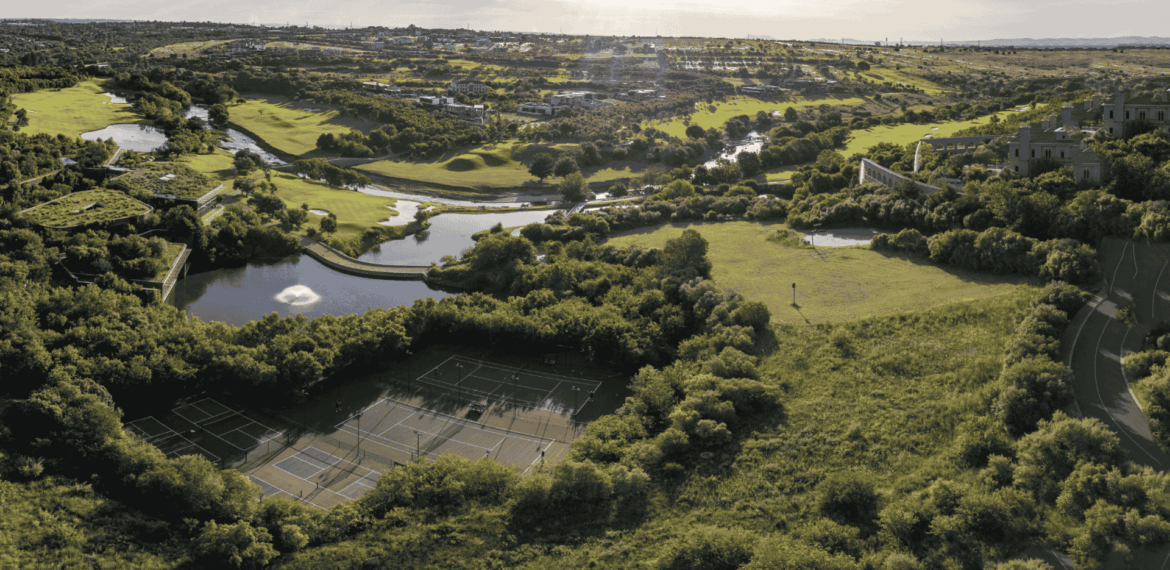Aerial view of a lush green landscape featuring tennis courts, winding rivers, ponds, open fields, and scattered trees under a bright sky.