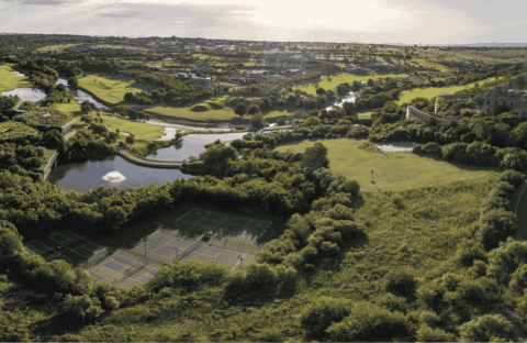 Aerial view of a lush green landscape featuring tennis courts, winding rivers, ponds, open fields, and scattered trees under a bright sky.