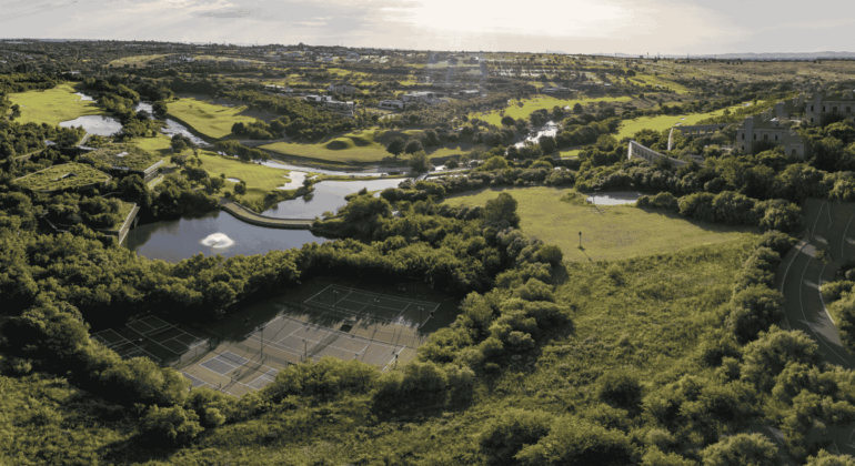 Aerial view of a lush green landscape featuring tennis courts, winding rivers, ponds, open fields, and scattered trees under a bright sky.