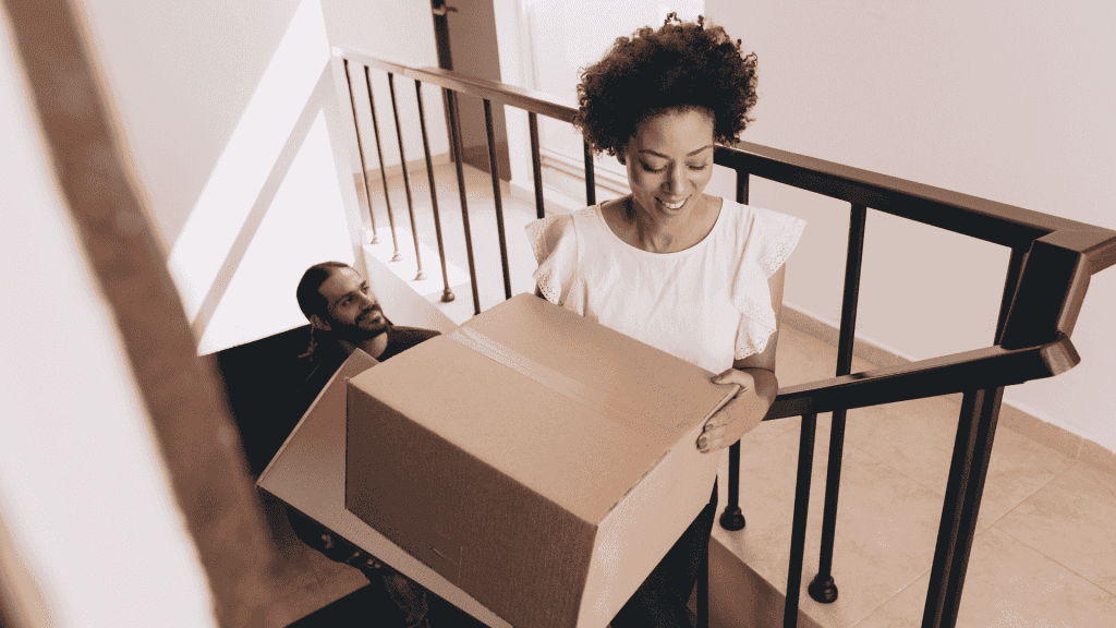 A woman smiles while carrying a large cardboard box up a staircase, followed by a man who is also climbing the stairs.