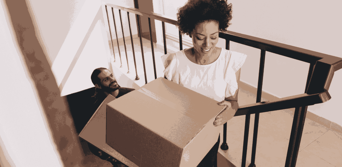 A woman smiles while carrying a large cardboard box up a staircase, followed by a man who is also climbing the stairs.