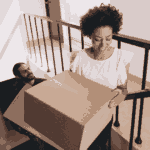 A woman smiles while carrying a large cardboard box up a staircase, followed by a man who is also climbing the stairs.
