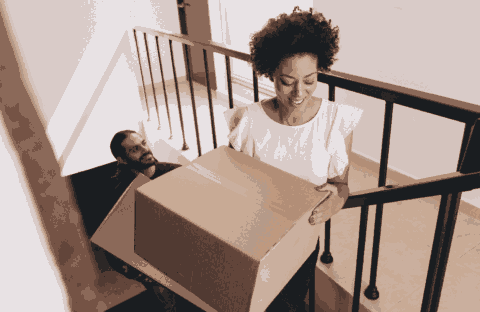 A woman smiles while carrying a large cardboard box up a staircase, followed by a man who is also climbing the stairs.