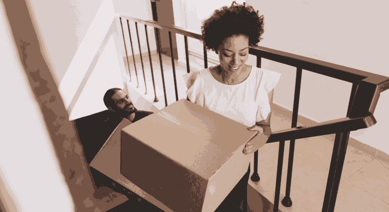 A woman smiles while carrying a large cardboard box up a staircase, followed by a man who is also climbing the stairs.