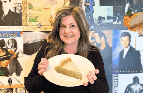 A woman smiles while holding a plate with a slice of pie in front of a wall covered with vintage music album covers. A floating cheesecake slice with caramel sauce and mint is visible in the top right corner.