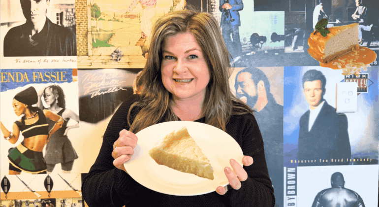 A woman smiles while holding a plate with a slice of pie in front of a wall covered with vintage music album covers. A floating cheesecake slice with caramel sauce and mint is visible in the top right corner.