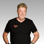 A smiling older man with light hair and a beard wears a black HOT 102.7FM T-shirt, a watch, and bracelets, standing against a plain gray gradient background.