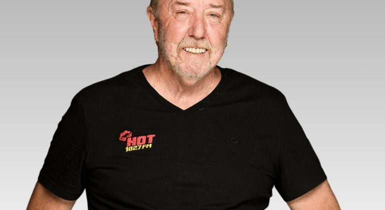 A smiling older man with light hair and a beard wears a black HOT 102.7FM T-shirt, a watch, and bracelets, standing against a plain gray gradient background.
