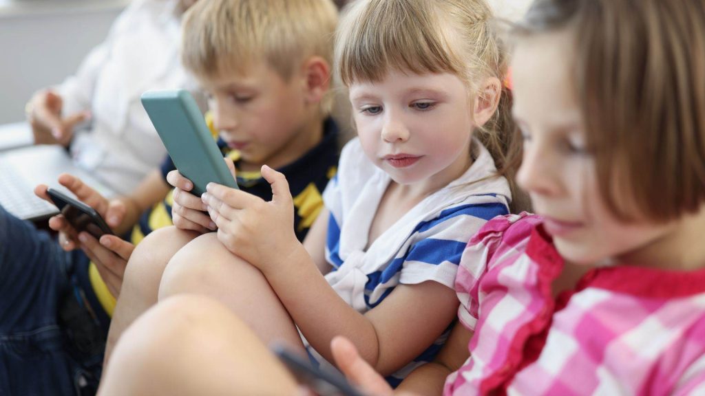Three young children sit closely together in a bright indoor setting, each focused on a smartphone or tablet screen. Dressed in casual striped clothing, they appear fully engaged with their devices, absorbed in their screen time.