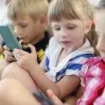 Three young children sit closely together in a bright indoor setting, each focused on a smartphone or tablet screen. Dressed in casual striped clothing, they appear fully engaged with their devices, absorbed in their screen time.