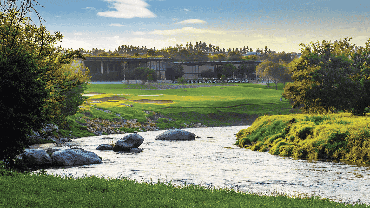 A scenic golf course with green fairways and sand bunkers, bordered by a gently flowing river and lush trees, under a partly cloudy sky at sunset.