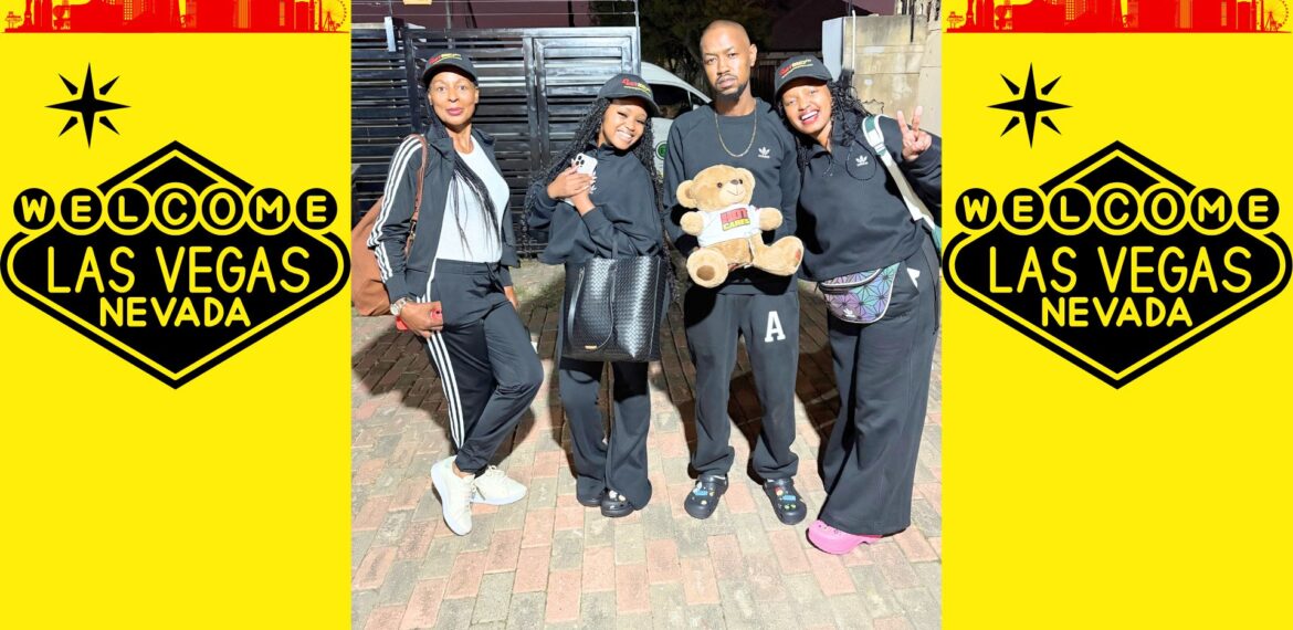 Four people in black outfits pose together, one holding a teddy bear, in front of a brick surface with Welcome to Las Vegas, Nevada signs on a bright yellow background on both sides.