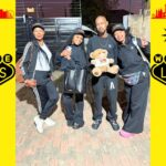 Four people in black outfits pose together, one holding a teddy bear, in front of a brick surface with Welcome to Las Vegas, Nevada signs on a bright yellow background on both sides.