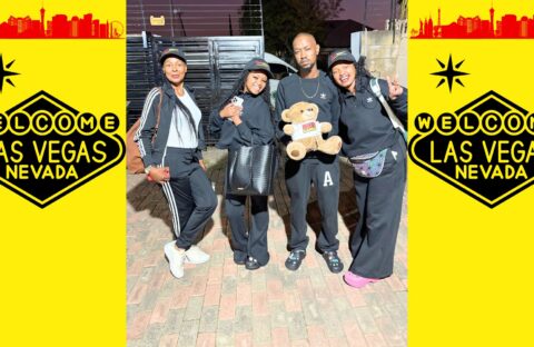Four people in black outfits pose together, one holding a teddy bear, in front of a brick surface with Welcome to Las Vegas, Nevada signs on a bright yellow background on both sides.