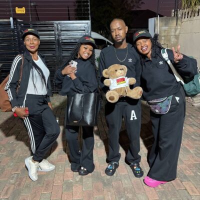 Four people in black Adidas tracksuits stand together outdoors on a brick surface. Three women smile, and a man in the middle holds a teddy bear. They all wear caps, and two women have bags. It is evening.