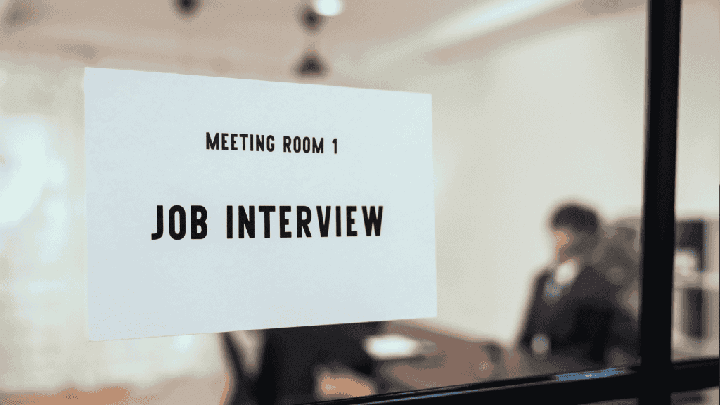 A sign reading Meeting Room 1 Job Interview is posted on a glass door. In the blurry background, two people are seated at a table, suggesting an interview is in progress.