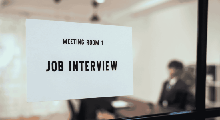 A sign reading Meeting Room 1 Job Interview is posted on a glass door. In the blurry background, two people are seated at a table, suggesting an interview is in progress.