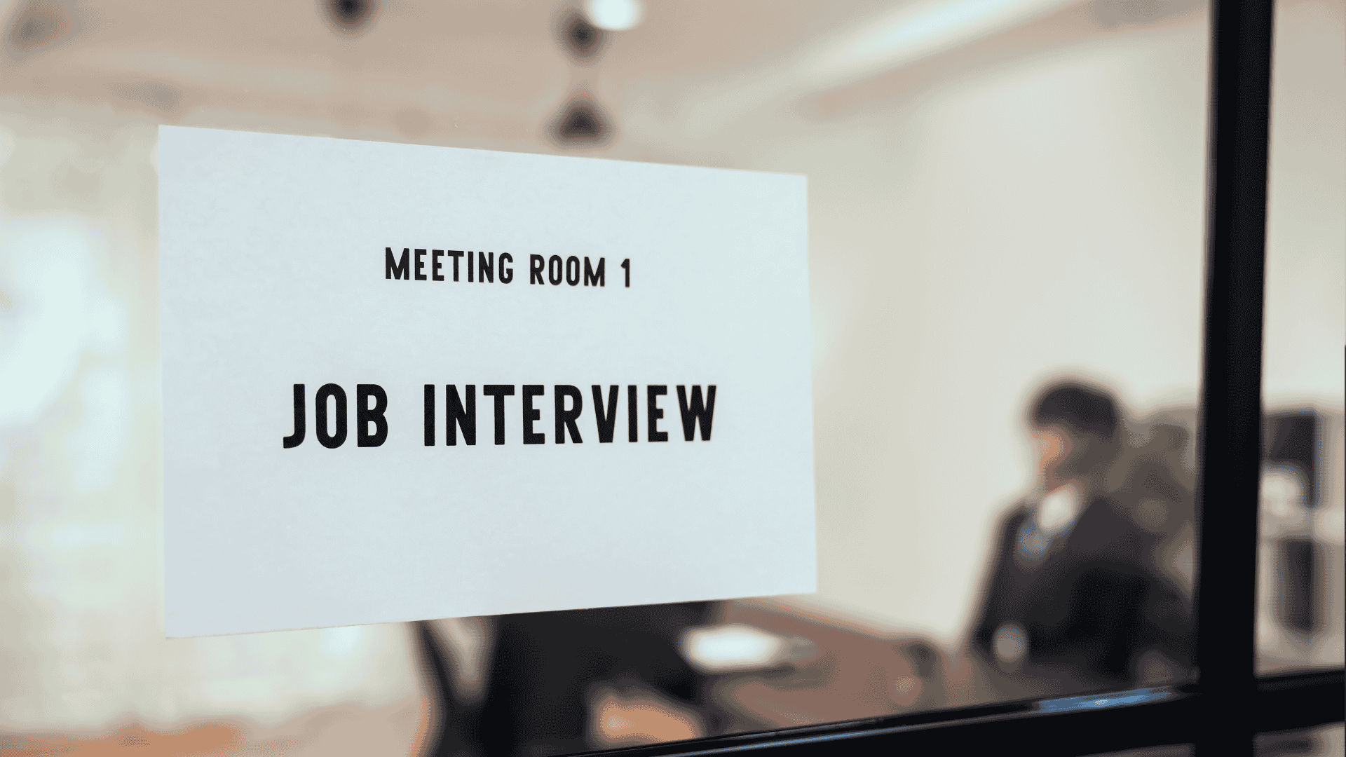 A sign reading Meeting Room 1 Job Interview is posted on a glass door. In the blurry background, two people are seated at a table, suggesting an interview is in progress.
