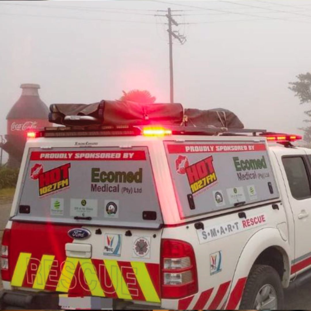 A white rescue vehicle with flashing red lights is parked in foggy conditions. The vehicle is covered with sponsor logos and RESCUE in yellow letters. A large Coca-Cola bottle sign is visible in the background.