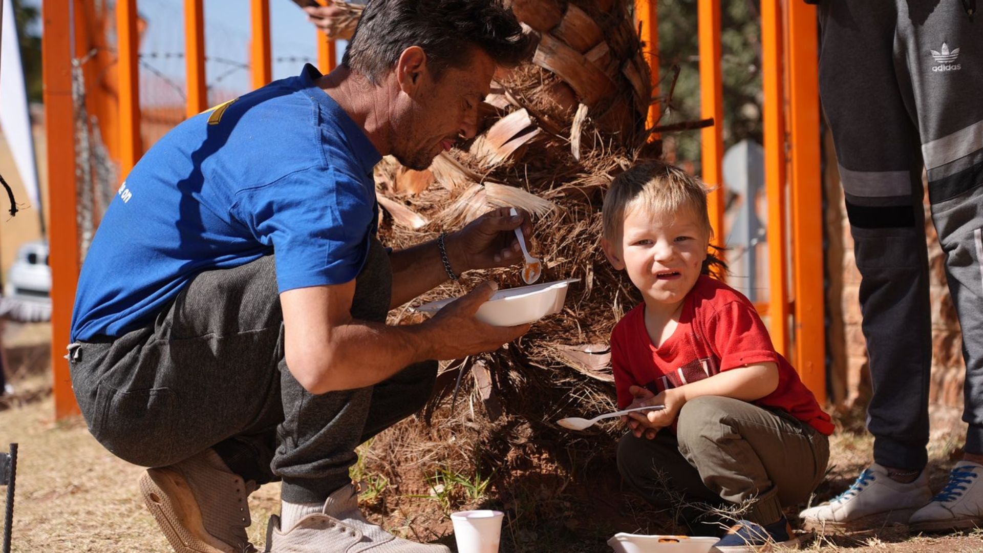 A man and a young boy crouch near a palm tree, eating food from bowls. The boy wears a red shirt and smiles at the camera, while the man in a blue shirt looks down at his bowl. Bright orange fence in the background.