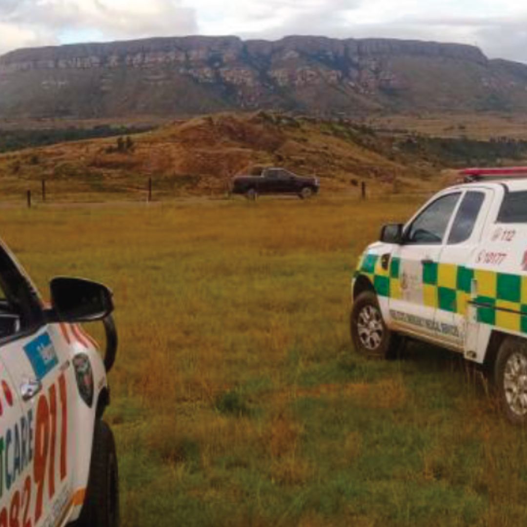 Two emergency response vehicles are parked on grassy terrain with a pickup truck in the background. A mountain range rises in the distance under a cloudy sky.