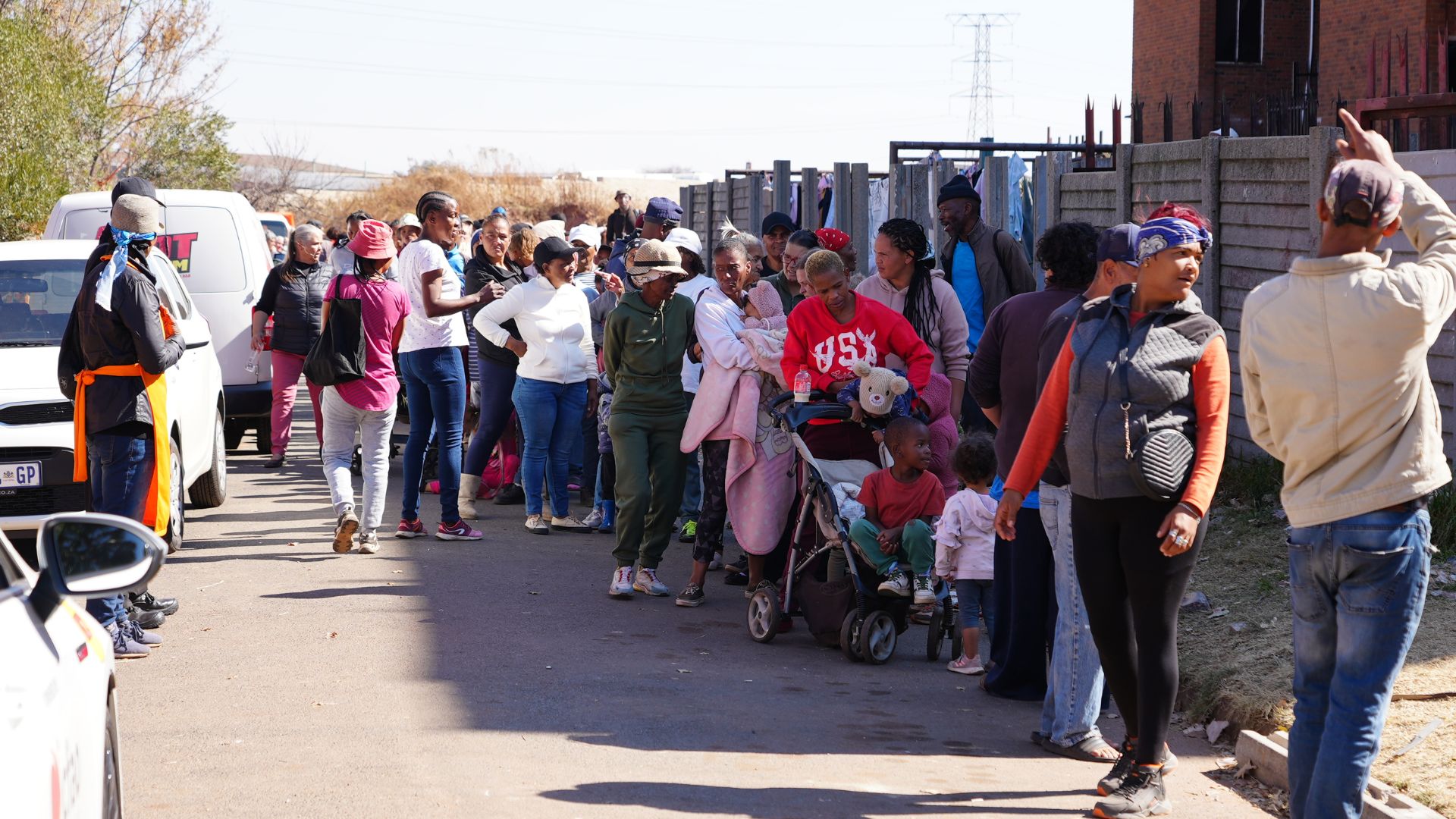 A long line of people, including adults and children, wait outdoors along a street. Some people stand, others have strollers, and a few are talking. The scene appears sunny and urban with buildings and vehicles nearby.