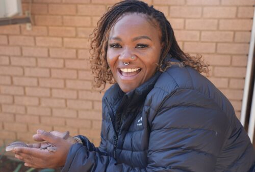 A smiling person with braided hair and a nose ring wears a black puffer jacket and holds a small lizard in their hands, standing in front of a brick wall.
