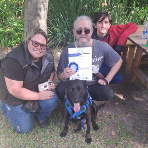 Three people and a black dog sit outdoors on grass. The man in the center holds a certificate and blue ribbon, while the dog sits in front wearing a harness. Trees and a wooden table are in the background. Everyone is smiling.