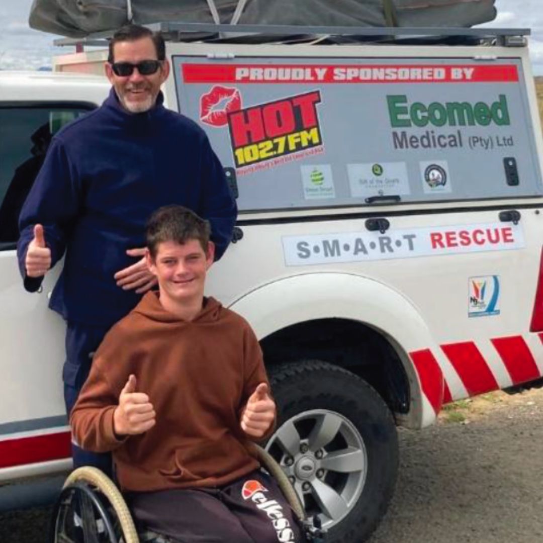 Two smiling people, one standing and one seated in a wheelchair, give thumbs up in front of a white rescue vehicle with sponsor logos including HOT 102.7 FM and Ecomed Medical.