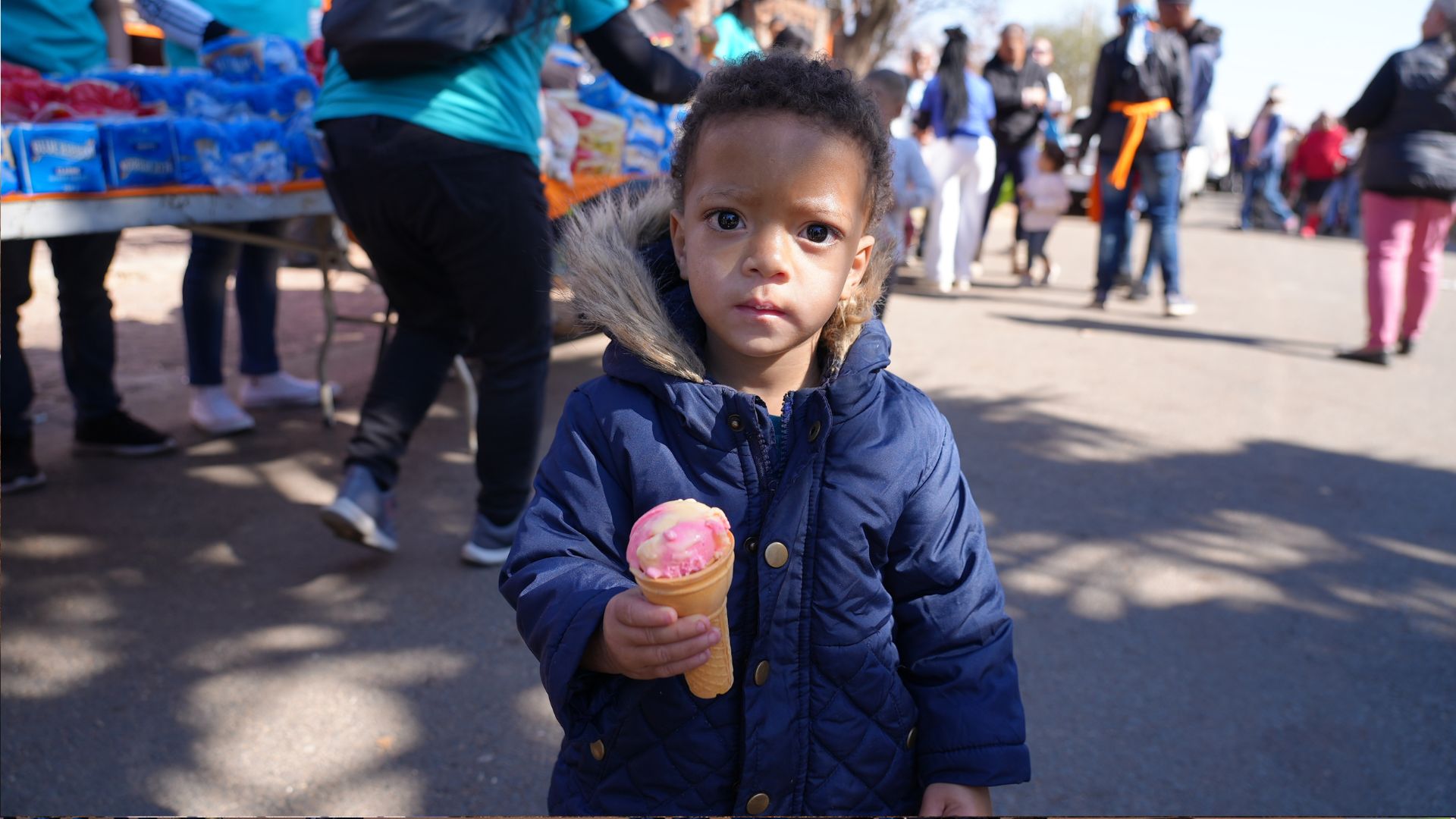 A young child in a navy blue coat holds an ice cream cone, looking at the camera. People and a food stall are visible in the background at an outdoor event.