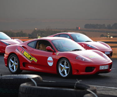 A red sports car with HOT 102.7 FM decals is parked on a racetrack, with another red sports car and a scenic landscape visible in the background. Tires are stacked in the foreground.