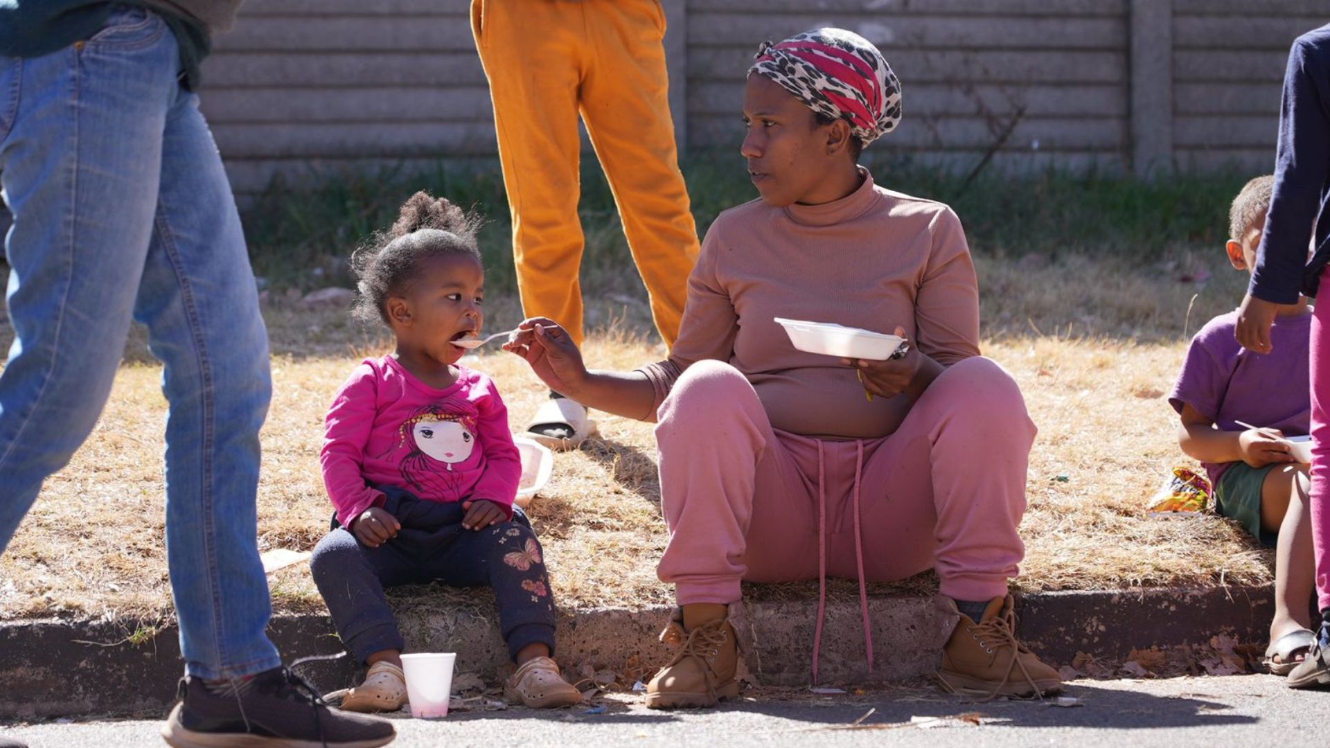 A woman sitting on a curb feeds a young girl with a spoon. The girl, dressed in a pink sweatshirt, sits next to her holding a cup. Other people stand or sit nearby, and a concrete fence is in the background.