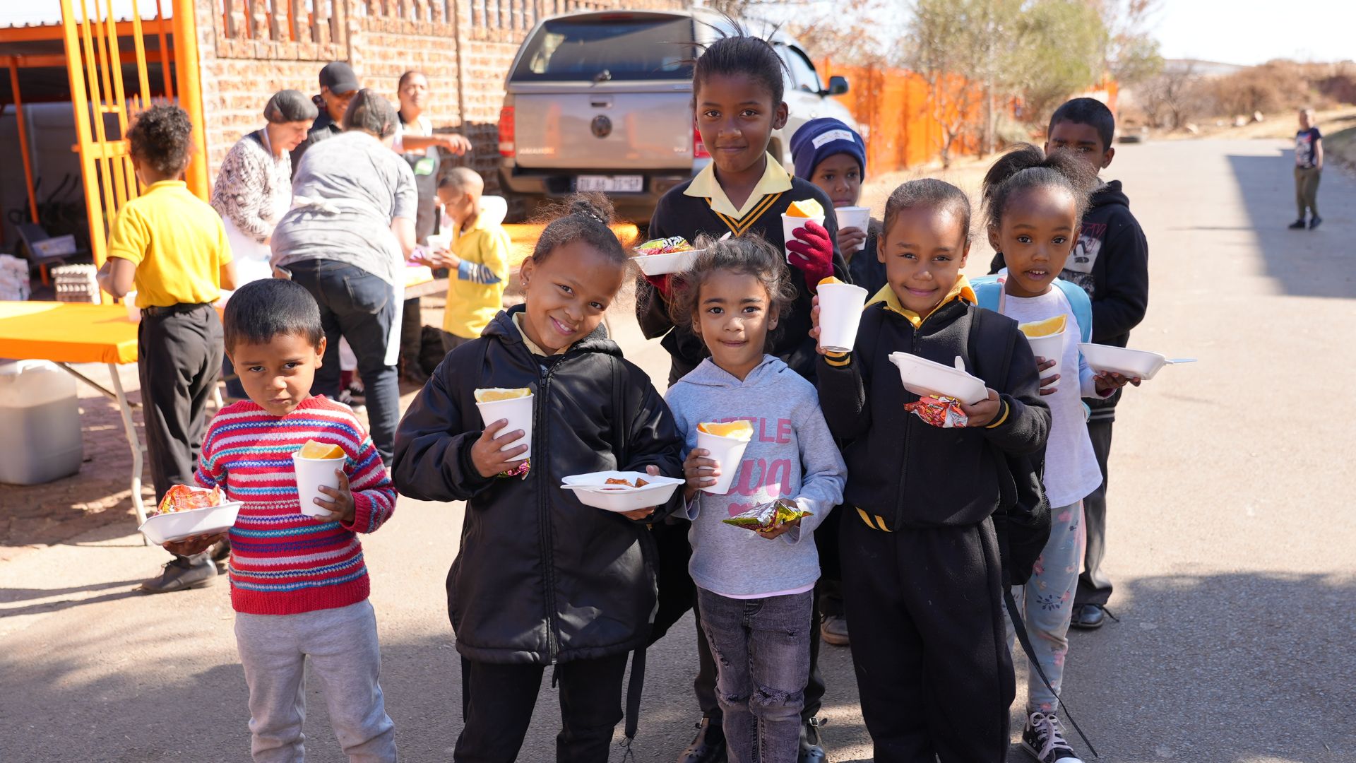 A group of smiling children stand outdoors in a line, holding plates and cups of food and snacks. Other people are serving food in the background near a parked car and orange fence on a sunny day.