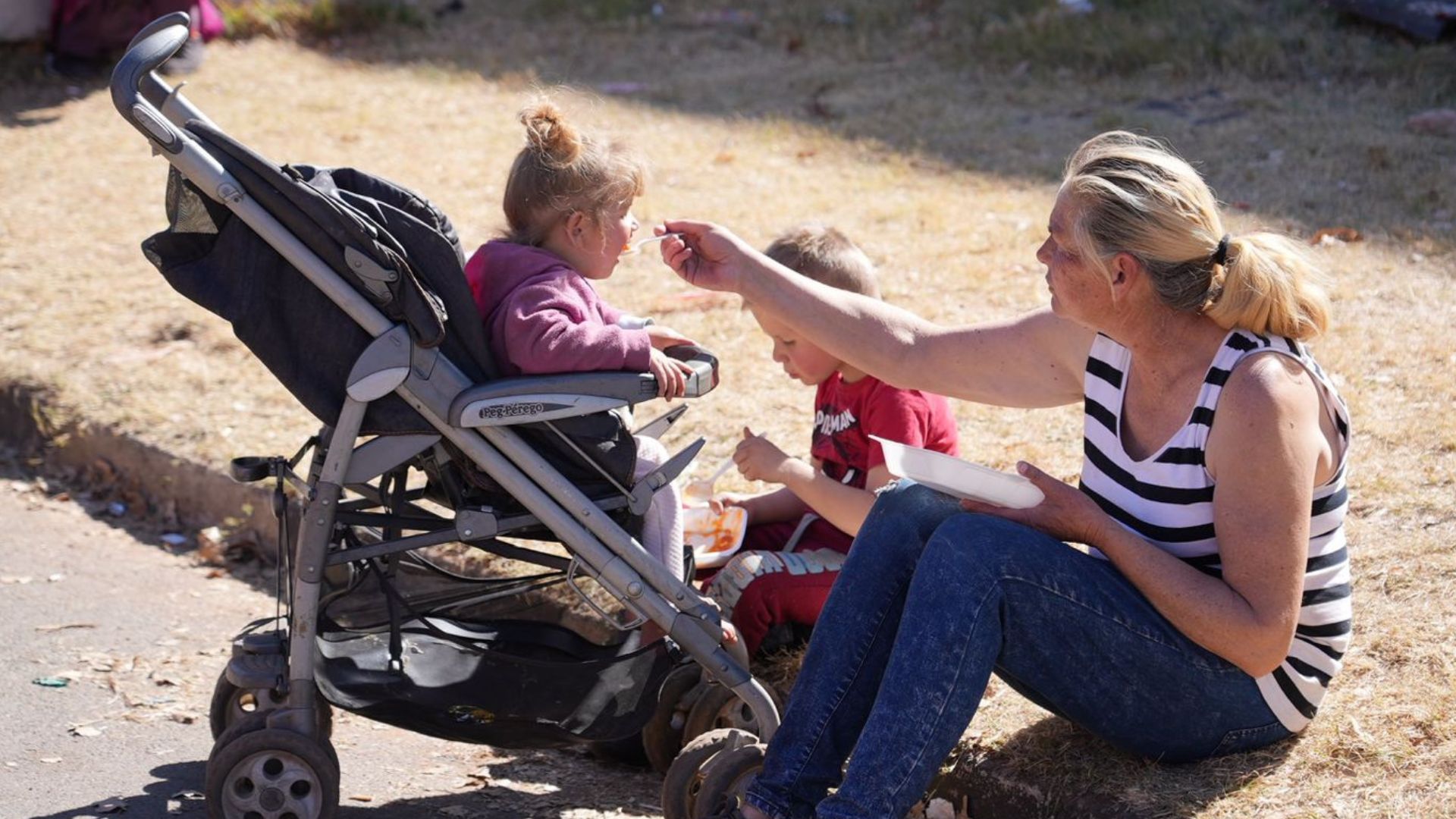 A woman in a striped tank top sits on the ground, feeding a young child in a stroller with a spoon. Another child sits nearby, eating from a plate. They are outdoors on a sunny day.