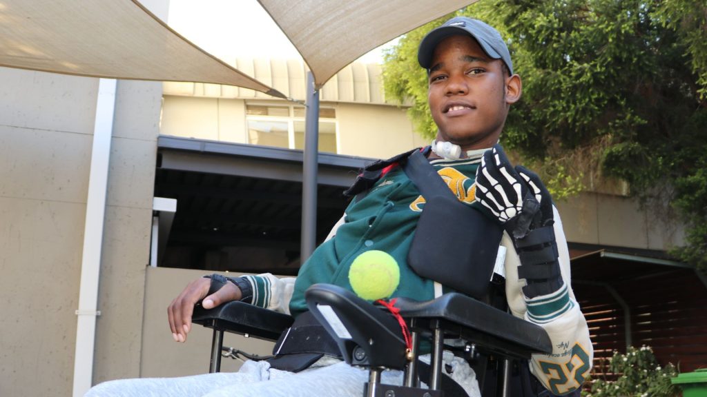 A young man in a wheelchair outdoors wears a cap, sports jacket, and a glove on one hand. He smiles and holds a tennis ball under his arm, with sunshades and a building visible in the background.